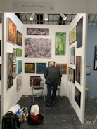 An art gallery booth displaying a variety of abstract paintings on white walls. A person with short hair and a dark jacket is standing in front of the artwork, closely observing them. The booth is organized with art pieces of different sizes and colors, and spotlights illuminate the space. On the floor, there are two folding chairs, a backpack, and a boxed item.