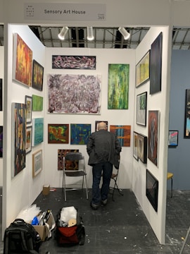 An art gallery booth displaying a variety of abstract paintings on white walls. A person with short hair and a dark jacket is standing in front of the artwork, closely observing them. The booth is organized with art pieces of different sizes and colors, and spotlights illuminate the space. On the floor, there are two folding chairs, a backpack, and a boxed item.
