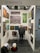 An art gallery booth displaying a variety of abstract paintings on white walls. A person with short hair and a dark jacket is standing in front of the artwork, closely observing them. The booth is organized with art pieces of different sizes and colors, and spotlights illuminate the space. On the floor, there are two folding chairs, a backpack, and a boxed item.