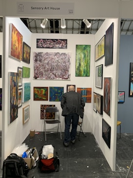 An art gallery booth displaying a variety of abstract paintings on white walls. A person with short hair and a dark jacket is standing in front of the artwork, closely observing them. The booth is organized with art pieces of different sizes and colors, and spotlights illuminate the space. On the floor, there are two folding chairs, a backpack, and a boxed item.
