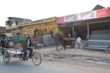 A busy street scene with a man riding a tricycle cart and a camel pulling a cart in front of multiple shops. The shops have colorful signage in Hindi and there are stacks of metal sheets and bricks in front. A tractor is visible, and several people are walking around, some in traditional attire.
