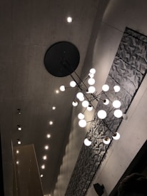 A modern ceiling design featuring a geometric light fixture with multiple spherical bulbs hanging from a central point. The ceiling is concrete, and there is a textured, abstract panel along one side, accompanied by recessed spotlights.