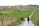 A person walks along a gravel path through a lush, expansive vineyard. The fields are neatly organized in rows on rolling hills, with a mix of green and brown patches indicating different stages of cultivation. Sparse trees and greenery dot the landscape under a clear, bright sky.