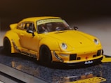 A yellow sports car with a sleek design and aerodynamic body, featuring prominent fender flares and a rear spoiler. The car has detailed features including side mirrors, headlights, and a visible windshield banner. It is placed on a reflective surface, enhancing the visual appeal.