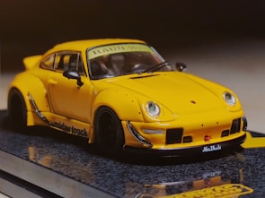 A yellow sports car with a sleek design and aerodynamic body, featuring prominent fender flares and a rear spoiler. The car has detailed features including side mirrors, headlights, and a visible windshield banner. It is placed on a reflective surface, enhancing the visual appeal.
