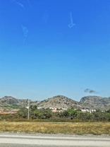 The image features a landscape with rolling hills in the background beneath a clear blue sky. Sparse vegetation is visible on the hills, while the foreground includes a cluster of houses surrounded by greenery with a stretch of dry grass beside a road.