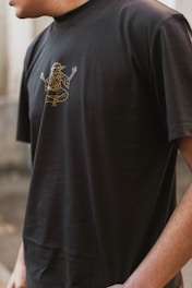 A person wearing a black t-shirt with a gold outline graphic of a mythological or spiritual figure in a meditative pose. The image captures the upper torso and partial arm of the individual in an outdoor setting.