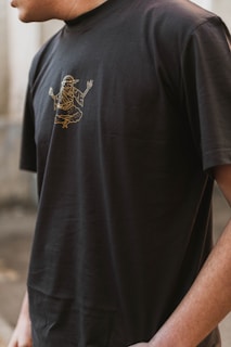 A person wearing a black t-shirt with a gold outline graphic of a mythological or spiritual figure in a meditative pose. The image captures the upper torso and partial arm of the individual in an outdoor setting.
