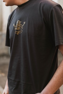 A person wearing a black t-shirt with a gold outline graphic of a mythological or spiritual figure in a meditative pose. The image captures the upper torso and partial arm of the individual in an outdoor setting.
