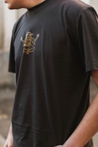 A person wearing a black t-shirt with a gold outline graphic of a mythological or spiritual figure in a meditative pose. The image captures the upper torso and partial arm of the individual in an outdoor setting.