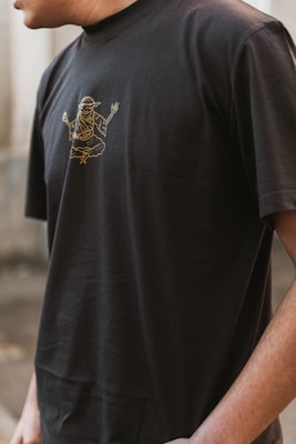 A person wearing a black t-shirt with a gold outline graphic of a mythological or spiritual figure in a meditative pose. The image captures the upper torso and partial arm of the individual in an outdoor setting.