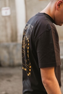 The image features a person wearing a black t-shirt with a graphic design on the back, which includes abstract patterns and checkerboard elements in yellow. The person stands in an outdoor setting with a blurred, neutral-toned background.