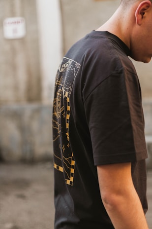 The image features a person wearing a black t-shirt with a graphic design on the back, which includes abstract patterns and checkerboard elements in yellow. The person stands in an outdoor setting with a blurred, neutral-toned background.