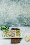 An assortment of elegant appetizers is displayed on a white platter placed on a wooden board. The appetizers include mini tarts filled with green mousse, cups topped with pomegranate seeds and microgreens, and delicate bites accented with herbs. A side dish of sliced cucumber and lime is set nearby, and a sliced lemon rests beside the serving board. The scene is set against a textured background with a net hanging on the wall, creating a subtle marine theme.