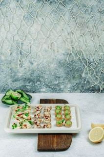 An assortment of elegant appetizers is displayed on a white platter placed on a wooden board. The appetizers include mini tarts filled with green mousse, cups topped with pomegranate seeds and microgreens, and delicate bites accented with herbs. A side dish of sliced cucumber and lime is set nearby, and a sliced lemon rests beside the serving board. The scene is set against a textured background with a net hanging on the wall, creating a subtle marine theme.