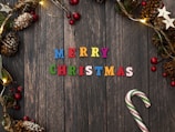 A festive arrangement featuring colorful letters spelling 'Merry Christmas' on a rustic wooden background. Surrounding the text are decorative elements such as pine cones, red and green berry clusters, glowing string lights, and white star ornaments. A candy cane with red and green stripes is placed on the right side.