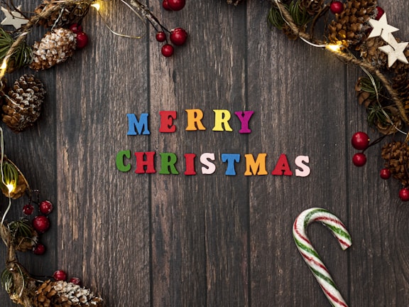 A festive arrangement featuring colorful letters spelling 'Merry Christmas' on a rustic wooden background. Surrounding the text are decorative elements such as pine cones, red and green berry clusters, glowing string lights, and white star ornaments. A candy cane with red and green stripes is placed on the right side.