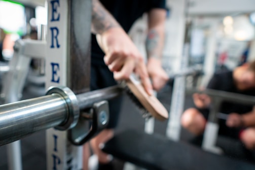 A person cleaning a metal barbell in a gym setting, using a brush to scrub the equipment. The gym environment is visible in the background, with various fitness equipment and another person out of focus.