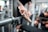 A person cleaning a metal barbell in a gym setting, using a brush to scrub the equipment. The gym environment is visible in the background, with various fitness equipment and another person out of focus.