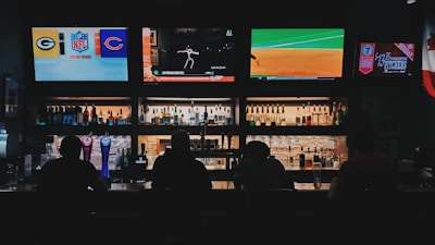 A bar counter with several people sitting in front of it is shown. Above the counter, there are multiple television screens displaying sports broadcasts, including football and baseball. Behind the counter, various bottles of alcohol are neatly arranged.