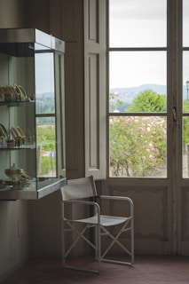 An elegant room corner with a large window looking out onto a lush green garden. A modern glass display case with golden decorative objects is mounted on the wall. A minimalist white metal chair is positioned near the window, receiving natural light.