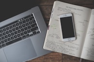 A close-up of a workspace featuring a laptop, an open notebook with handwritten notes, and a smartphone. The laptop is grey and positioned to the left, while the notebook and smartphone are placed on the right. The notes cover a range of topics, with visible diagrams and text in different colors.