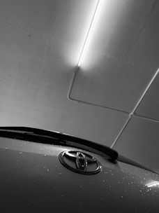 A black and white image featuring the front portion of a car with a visible Toyota emblem. Water droplets are scattered across the vehicle's surface, suggesting it's recently been in rain or washed. Above, fluorescent lighting lines the ceiling of an indoor parking setting, casting a stark glow on the scene.