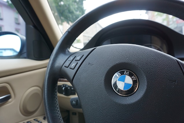 A close-up view of a car's steering wheel featuring the BMW logo at the center, with control buttons visible on the steering column. The interior is beige, and the side mirror reflects part of the exterior environment. Trees and buildings can be seen through the windshield.