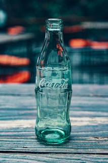 An empty, classic Coca-Cola glass bottle is positioned upright on a wooden surface. The background is blurred, focusing attention on the bottle's greenish tint and iconic branding.