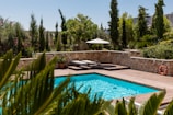 A serene outdoor pool area surrounded by lush greenery and tall trees. The scene features a rectangular swimming pool with clear blue water, wooden decking, and sun loungers under a white umbrella. Natural stone walls enclose the space, and there is a bright orange lifebuoy attached to one wall. Various potted plants and vibrant flowers enhance the tranquil setting.