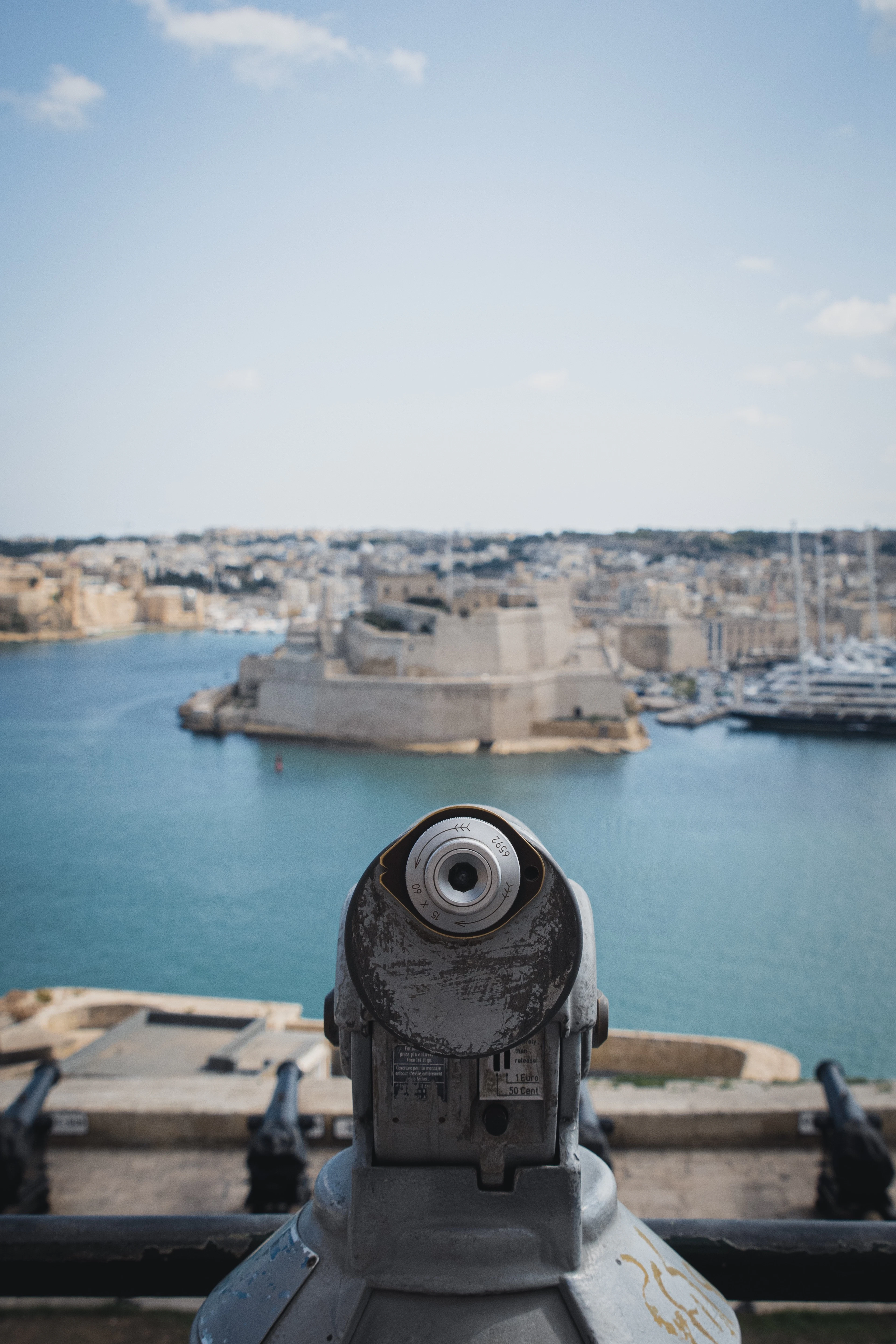 A weathered coin-operated tower viewer on the terrace above Valletta's Saluting Battery, aimed across the blue Grand Harbour at the low stone bastions of Fort St Angelo and the Three Cities beyond.