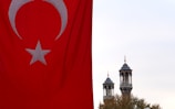 A large Turkish flag with a prominent red background and a white star and crescent is prominently displayed on the left side. In the background, two ornate minarets rise against a cloudy sky, partially obscured by treetops.