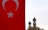 A large Turkish flag with a prominent red background and a white star and crescent is prominently displayed on the left side. In the background, two ornate minarets rise against a cloudy sky, partially obscured by treetops.