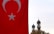 A large Turkish flag with a prominent red background and a white star and crescent is prominently displayed on the left side. In the background, two ornate minarets rise against a cloudy sky, partially obscured by treetops.