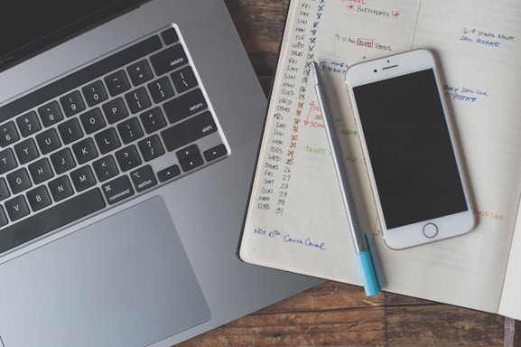 A laptop keyboard is visible alongside an open notebook filled with handwritten notes and schedules. A smartphone with a black screen rests atop the notebook, and a pen is placed next to it. The notebook pages contain a calendar layout with various tasks and reminders written in different colors.