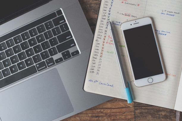 A laptop keyboard is visible alongside an open notebook filled with handwritten notes and schedules. A smartphone with a black screen rests atop the notebook, and a pen is placed next to it. The notebook pages contain a calendar layout with various tasks and reminders written in different colors.