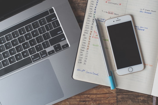 A laptop keyboard is visible alongside an open notebook filled with handwritten notes and schedules. A smartphone with a black screen rests atop the notebook, and a pen is placed next to it. The notebook pages contain a calendar layout with various tasks and reminders written in different colors.