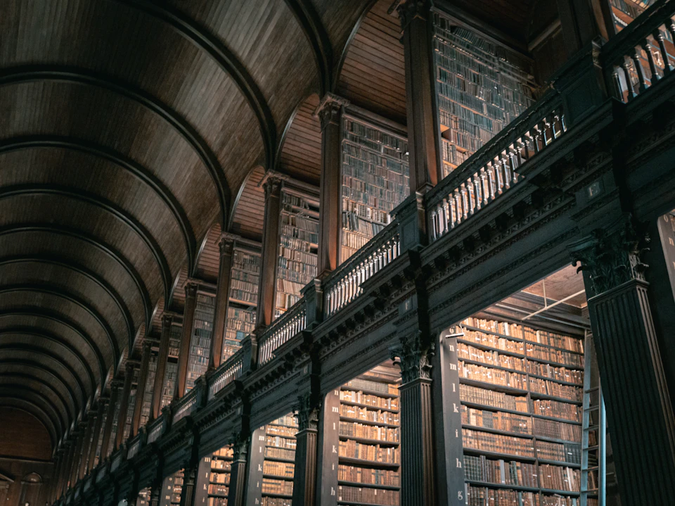 The image depicts a grand, old library with a high ceiling and wooden beams that add to its rustic charm. The perspective is from below, looking up at the rows of bookshelves filled with numerous books. The lighting in the room is dim, creating an atmosphere of quiet contemplation and intellectual pursuit.