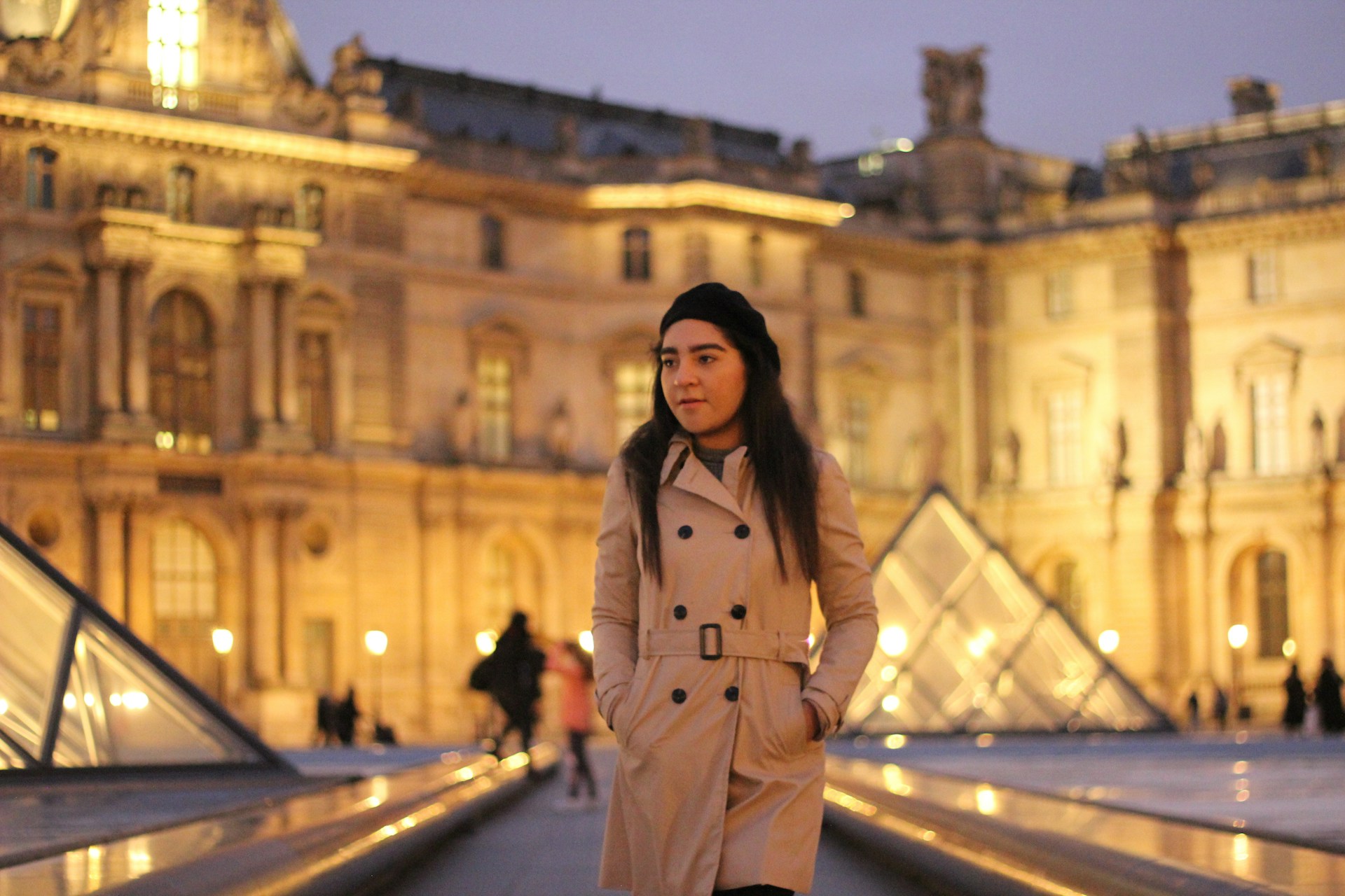 A stylish woman in a soft beige trench coat standing on a sunlit city street with neutral-toned buildings in the background.