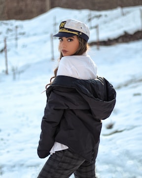 A person wearing a navy captain's hat and a black padded jacket stands outdoors, looking over their shoulder. The background is covered in snow with a hint of fencing visible, suggesting a cold, winter setting. The person has long hair and is dressed warmly, indicating winter attire.