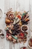 A richly arranged charcuterie board featuring a variety of cheeses, fresh fruits like figs, grapes, and pears, along with crackers, nuts, and olives. Small bowls contain black and green olives, and there are decorative herbs and spreads interspersed. The arrangement rests on a rustic wooden surface with a rustic knife with a turquoise handle included.