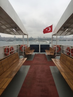 A ferry with wooden benches and lifebuoys on both sides is sailing on a body of water, featuring a red Turkish flag with a white star and crescent. The cityscape is visible in the background under a cloudy sky.