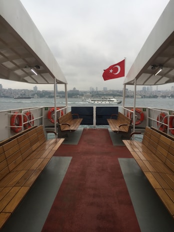 A ferry with wooden benches and lifebuoys on both sides is sailing on a body of water, featuring a red Turkish flag with a white star and crescent. The cityscape is visible in the background under a cloudy sky.