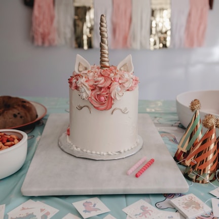 A unicorn-themed cake decorated with pink and white flowers, a golden horn, and ears sits on a table. Nearby, there are colorful party hats with gold tassels, bowls of snacks, and small printed pictures featuring unicorns. The background includes decorative tassels in pink, white, and metallic gold.