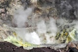A rocky landscape dominated by a crater that is emitting large amounts of steam and gas. The foreground consists of dark rocks, transitioning into lighter shades with hints of yellow and green near the gas emissions. A group of people stand at the edge of the crater, providing a sense of scale against the rugged terrain.