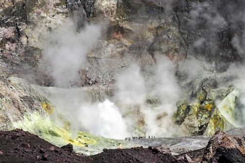 A rocky landscape dominated by a crater that is emitting large amounts of steam and gas. The foreground consists of dark rocks, transitioning into lighter shades with hints of yellow and green near the gas emissions. A group of people stand at the edge of the crater, providing a sense of scale against the rugged terrain.
