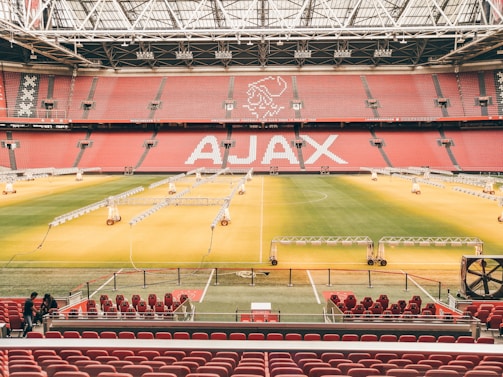 A large football stadium with red seating and a well-maintained green field. The word 'AJAX' is prominently displayed on the seats. The stadium is equipped with lighting rigs and a partly open roof structure.