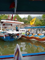 Several colorful boats are moored on a calm body of water, surrounded by lush greenery. The boats have vivid designs and a yellow flag is visible. A person wearing a blue shirt stands on one of the boats.