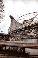 A futuristic bridge with a complex metallic structure spans over a body of water. In the background, a modern architectural building with a unique ship-like design at its top towers above the other structures.