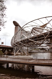 A futuristic bridge with a complex metallic structure spans over a body of water. In the background, a modern architectural building with a unique ship-like design at its top towers above the other structures.